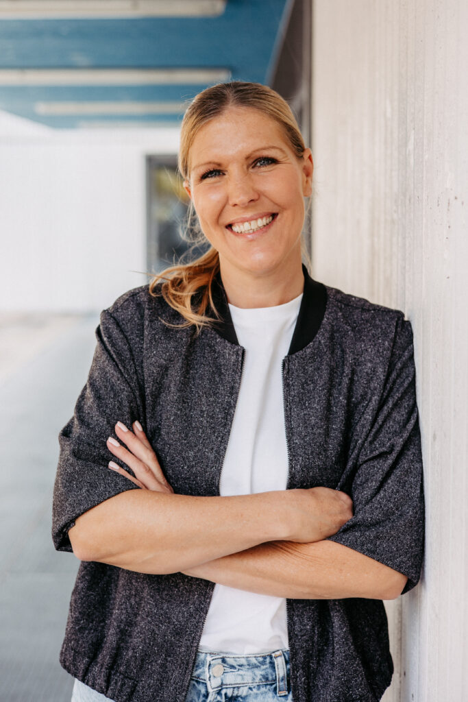 Bewerbungsfoto, Bewerbungsportrait, Bewerbungsbild. Outdoor an einem Gebäude aufgenommen. Person lehnt locker an einer Steinwand, hat die Arme verschränkt und schaut freundlich in die Kamera