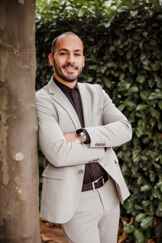 Lockeres Businessshooting. Outdoor. Mann lehnt an einem Baumstamm, hat die Arme verschränkt und lächelt in die Kamera. So kann ein Berufs-Portrait aussehen.