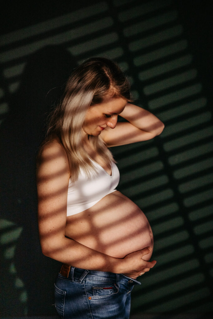 Babybauchshooting im Studio mit künstlichem Sonnenlicht, das aussieht als scheint es durch eine Jalousie. Minimalistische Fotografie
