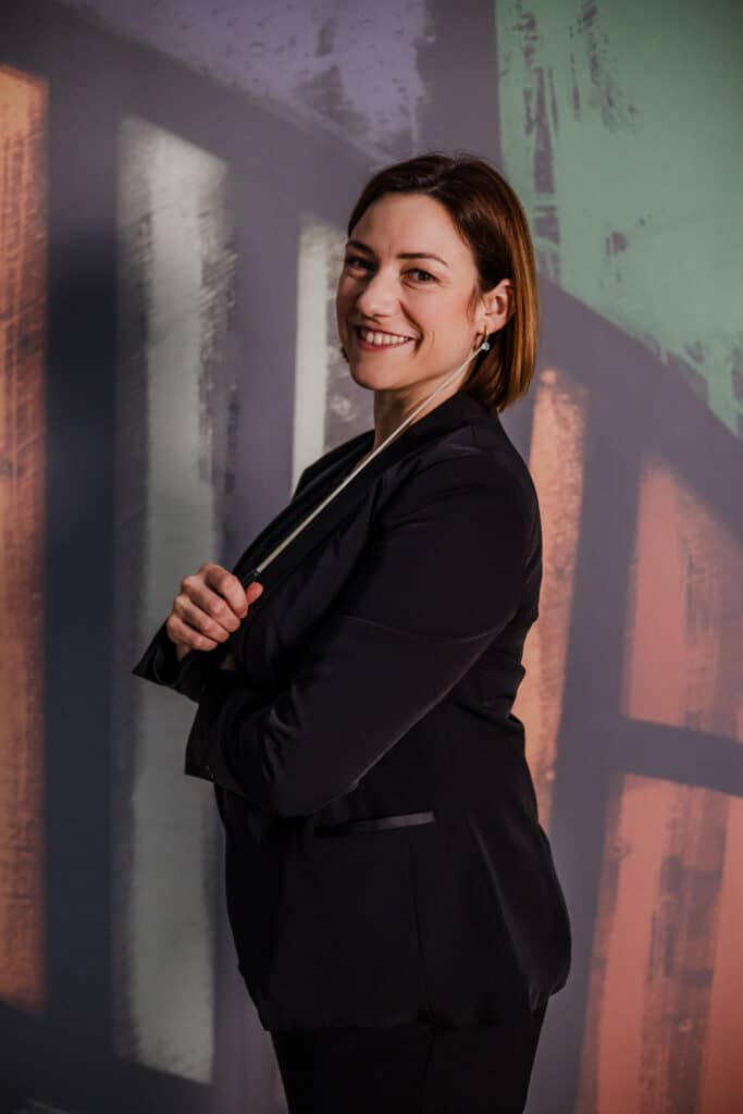 Die Chefin im Fokus. Portrait der Geschäftsführerin. Eine Dirigentin mit Taktstock vor einer bunten Wand. Keine langweiligen Bilder